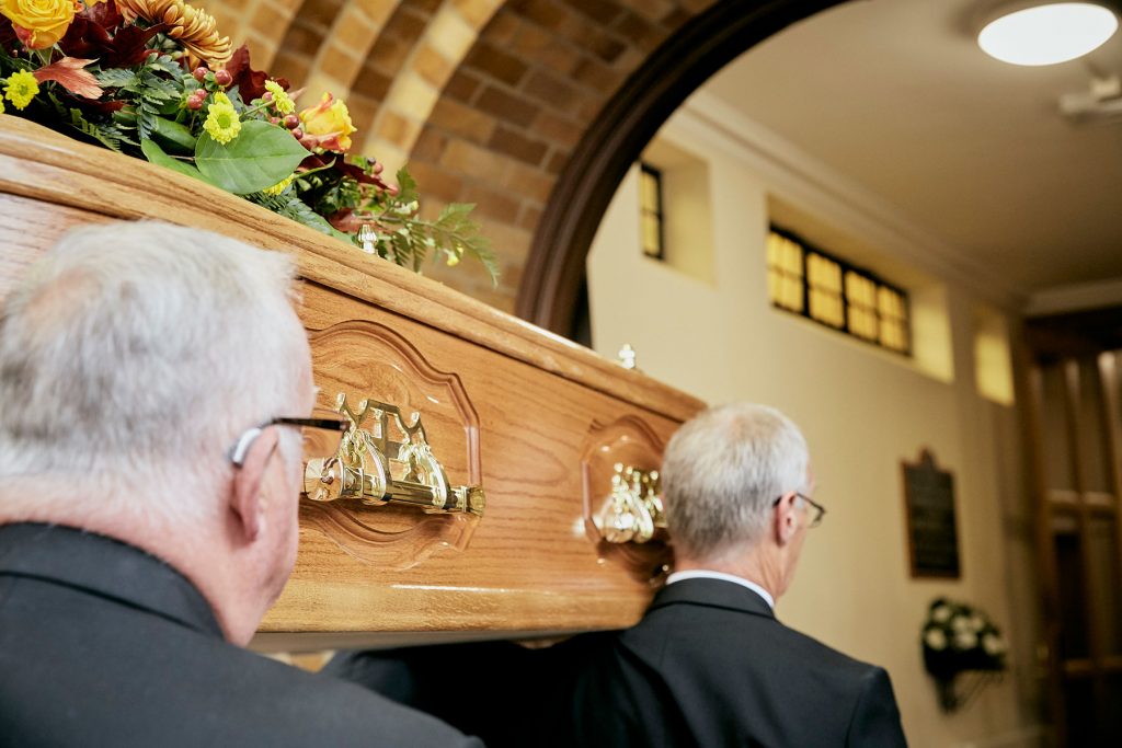 Two pallbearers carrying a wooden coffin with a floral arrangement on top